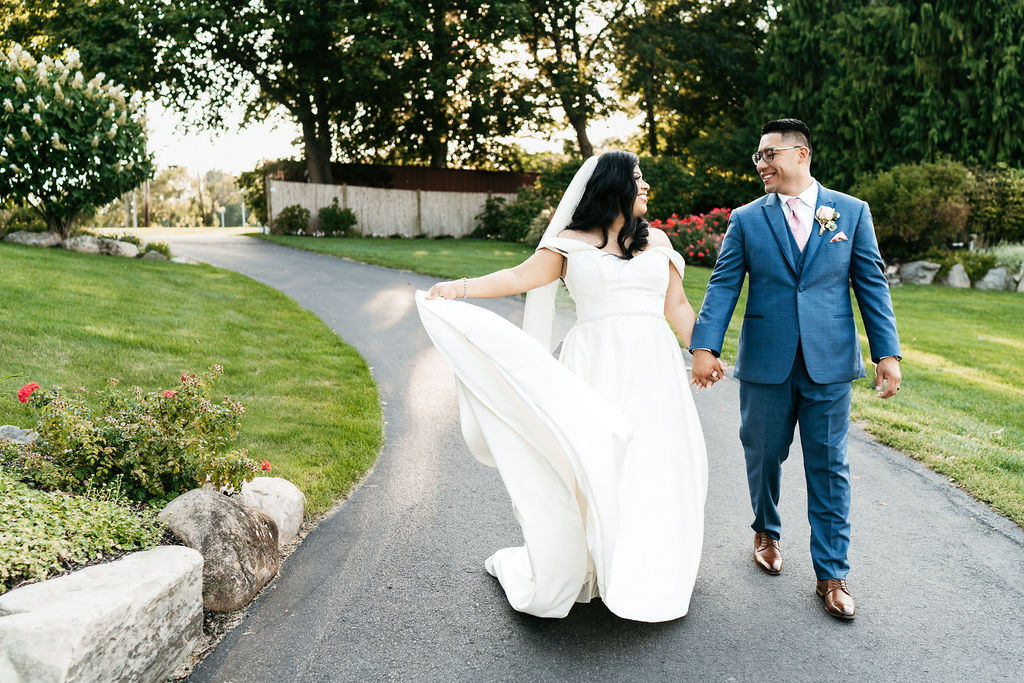 A bride and groom are smiling at one another and holding hands. The bride is twirling the skirt of her dress as they walk down a paved road at their venue. Flowers bloom in the background at their sweet summer wedding.