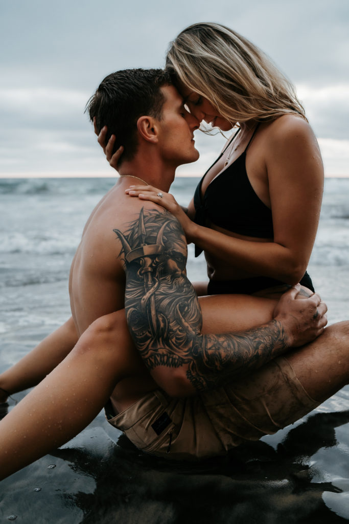 A tattooed couple is leaning their foreheads together while sitting on the Torrey Pines shore. The sand looks black and the waves are rolling gently behind them. 